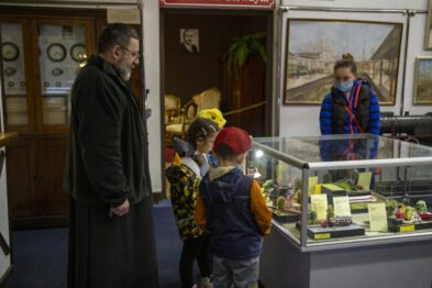 Mężczyzna w ciemnym ubraniu stoi obok grupy dzieci i wskazuje na eksponaty umieszczone wewnątrz szklanej witryny na wystawie poświęconej satyrom kolejowym. W tle znajduje się kolejna vitryna oraz obrazy związane z kolejnictwem. Dzieci skupione są na oglądanych przedmiotach, mają na sobie kolorowe ubrania.