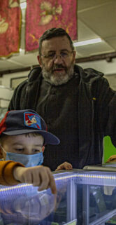 Grupa osób skupia się na oglądaniu eksponatów prezentowanych w szklanych witrynach. Osoba dorosła, prawdopodobnie przewodnik, wskazuje na jeden z przedmiotów, angażując uwagę dwójki młodszych uczestników wydarzenia. W tle zauważalna jest zawieszona dekoracja oraz kolejna witryna z eksponatami.