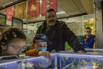 Grupa osób skupia się na oglądaniu eksponatów prezentowanych w szklanych witrynach. Osoba dorosła, prawdopodobnie przewodnik, wskazuje na jeden z przedmiotów, angażując uwagę dwójki młodszych uczestników wydarzenia. W tle zauważalna jest zawieszona dekoracja oraz kolejna witryna z eksponatami.