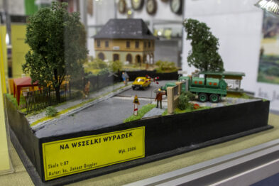 Model kolejowy przedstawia scenę z miniaturową zieloną lokomotywą parową i dwoma wagonami na torach, otoczonymi przez drzewa i trawy. Obok torów widoczny jest żółty samochód oraz budynek stacji kolejowej z ciemnym dachem. Na przedzie makiety umieszczono etykietę z napisem 