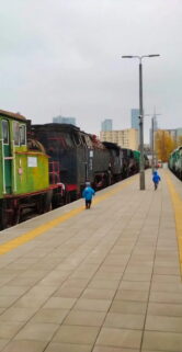 Dzieci bawią się na peronie kolejowym, gdzie obok siebie ustawione są różne lokomotywy w barwach zielonych i czarnych. Chodnik peronu wyłożony jest płytami, a w tle widnieje wysoka zabudowa miejska. Pogoda jest pochmurna, a peron oświetlony dziennym światłem.