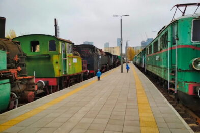 Dzieci bawią się na peronie kolejowym, gdzie obok siebie ustawione są różne lokomotywy w barwach zielonych i czarnych. Chodnik peronu wyłożony jest płytami, a w tle widnieje wysoka zabudowa miejska. Pogoda jest pochmurna, a peron oświetlony dziennym światłem.