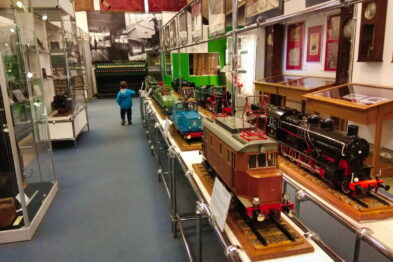 W muzealnej sali wystawione są modele kolejek i lokomotyw, ustawione na wysokich postumentach. Rodzina zwiedza ekspozycję – dorosły i dziecko idą środkiem między eksponatami. Ściany są udekorowane zdjęciami i pamiątkami kolejnictwa.