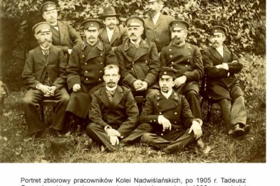 Grupa mężczyzn w historycznych mundurach kolejarskich pozują do wspólnego zdjęcia w plenerze. Wszyscy mają na sobie czapki z daszkami i są ubrani w ciemne garnitury z wyraźnymi insygniami. Na środku zdjęcia siedzi starszy mężczyzna w jasnym mundurze z odznaczeniami, otoczony przez pozostałych pracowników kolei.
