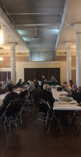 W przestronnej sali z kolumnami i lustrzanie odbijającymi wnętrze zgromadzili się uczestnicy spotkania siedzący przy długich stołach. Osoby są zwrócone twarzami w kierunku mówcy umieszczonego na przeciwległym końcu sali. W centralnej części fotografii widać białe flagi z emblematami, które są rozmieszczone pomiędzy stołami.