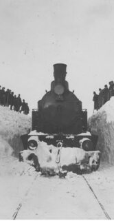 Steam locomotive is visible at the center of the image, surrounded by snow and outlined by snow-cleared tracks. A group of people stands to the left of the locomotive, partially elevated on the snowbank. The sky is overcast, and the photo conveys a historical ambiance due to its monochromatic nature.