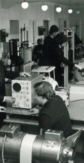 Na fotografii widać cztery osoby w pomieszczeniu pełnym sprzętu technicznego; dwóch mężczyzn pracuje przy dużym, okrągłym urządzeniu mechanicznym, zaś pozostali dwaj zajmują się obsługą urządzeń elektronicznych. Każdy z nich nosi ciemny strój, a w tle widać różnego rodzaju przyrządy i wyposażenie, które sugeruje warsztat lub laboratorium techniczne. Pomieszczenie jest jasne i uporządkowane, a skupienie pracowników na wykony