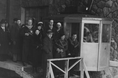 Grupa osób stoi obok małej kabiny kolejki linowej, która jest umieszczona na konstrukcji wspierającej. Osoby są ubrane w ciemne, epokowe ubrania, z których wiele nosi kapelusze. Kabina kolejki jest otwarta, a tło za grupą tworzy kamienna ściana, będąca prawdopodobnie częścią stacji.