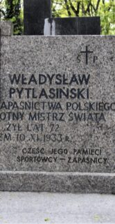 Nagrobek z wyrytym imieniem i nazwiskiem, datami życia oraz tytułami "Mistrz Olimpijski, Polskiego, Wielokrotny Mistrz Świata", jest usytuowany na cmentarzu. Kwiaty są umieszczone na i obok pomnika. Tło stanowią inne nagrobki i drzewa, co sugeruje spokojną, zalesioną część cmentarza.