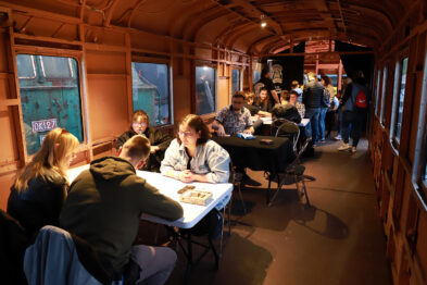 W historycznym wnętrzu wagonu kolejowego siedzą ludzie przy rozłożonych na stolikach planszach gier. Światło wpada przez okna wagonu, tworząc przyjemną atmosferę. Po lewej stronie zdjęcia widać przechadzających się ludzi oglądających ekspozycję.