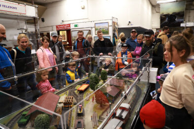 Grupa ludzi z różnych grup wiekowych zgromadziła się wokół makiet kolejowych znajdujących się pod szklanymi osłonami. Dzieci i dorośli obserwują z zainteresowaniem miniaturowe modele pociągów oraz detalicznie wykonane elementy krajobrazu kolejowego. Przewodnicy i pracownicy muzeum w czerwonych czapkach wspierają zwiedzających, odpowiadając na pytania i opowiadając o eksponatach.