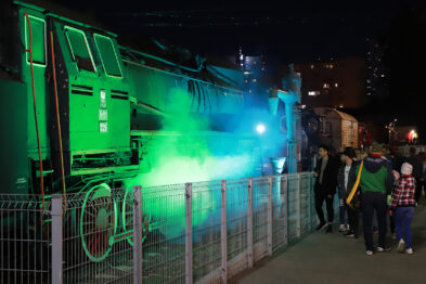 Ludzie zgromadzili się wokół zielono podświetlonego parowozu, który jest wyeksponowany na zewnątrz. W tle szeregi innych zabytkowych pociągów są widoczne, a scenerię dopełnia nocne oświetlenie. Atmosfera wydarzenia jest uchwycenia dzięki ciepłym kolorom świateł i zainteresowaniu uczestników stojących przy ogrodzeniu.