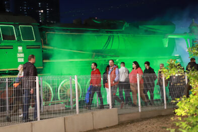 Grupa ludzi stoi przy zielono podświetlonym lokomotywowi na zewnątrz w nocy, wykonując czynności związane z eksploracją eksponatu. Lokomotywa jest oświetlona z dołu, tworząc widowiskowe efekty świetlne, które podkreślają jej kontury i szczegóły konstrukcyjne. Wokół grupy i lokomotywy widać ogrodzenie i inne elementy infrastruktury kolejowej.
