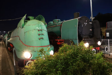 Zielona lokomotywa parowa z czerwonymi obramówkami stoi na torach, oświetlona w nocy, co podkreśla jej kształty i szczegóły konstrukcji. Za nią znajduje się ciemniejsza lokomotywa bez wyraźnych oznaczeń. Oświetlenie punktowe i linie kolejowe tworzą urokliwy krajobraz techniczny, podkreślający charakter muzealnej wystawy kolejnictwa.