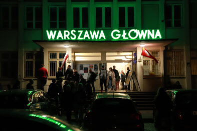 Grupa ludzi gromadzi się przed budynkiem z zielono podświetlonym napisem 