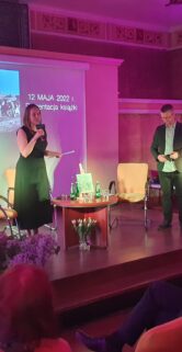 Jest to wnętrze eleganckiego pomieszczenia, gdzie odbywa się wydarzenie literackie. Na pierwszym planie widać siedzących ludzi skupionych na prezentacji. Na scenie stoi kobieta przy mikrofonie i mężczyzna w ciemnym stroju, zajął miejsce na fotelu; w tle znajdują się ekrany z projekcją, a dekoracje kwiatowe dodają uroczystego charakteru wydarzeniu.