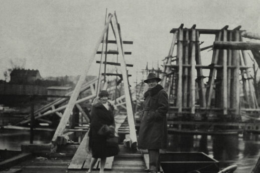Dwie osoby w ciemnych płaszczach i kapeluszach stoją na drewnianym pomostowym moście. W tle widać konstrukcję kolejowego mostu z widocznymi elementami drewnianymi i stalowymi. Otoczenie jest mgliste, co nadaje zdjęciu historyczny charakter.