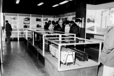 Ekspozycja muzealna zawiera modele lokomotyw oraz tablice informacyjne przedstawiające historię kolei; odwiedzający przeglądają wystawę, co świadczy o interakcji publiczności z eksponatami. Jest to czarno-białe zdjęcie wnętrza muzeum z grupą ludzi skupionych na oglądaniu prezentowanych obiektów.