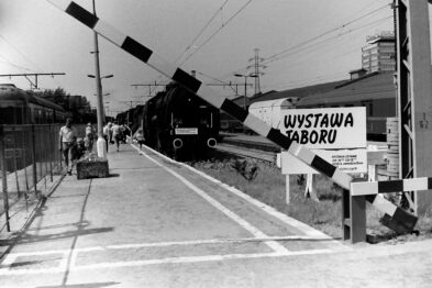 Czarno-biała fotografia przedstawia ekspozycję zabytkowego taboru kolejowego ze znakiem informującym o wystawie. Lokomotywa parowa i wagony są wyeksponowane na zewnątrz na torach kolejowych. W tle znajdują się budynki dworcowe i infrastruktura kolejowa.