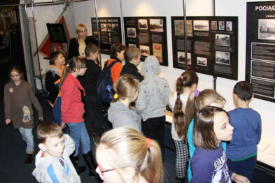 Grupa dzieci w wieku szkolnym zwiedza wystawę muzealną; oglądają eksponaty i informacje prezentowane na ścianie. Przewodnik lub nauczyciel stoi z boku, obserwując grupę. Wnętrze jest dobrze oświetlone, a na zdjęciu dominuje kolorowa odzież zwiedzających.