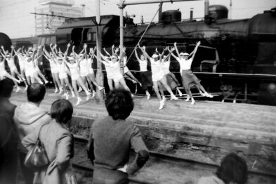 Grupa gimnastyków wykonuje pokaz na zewnątrz; w tle stoi parowóz i wagony kolejowe. Widzowie obserwują występ, stojąc za drewnianym ogrodzeniem. Całość ma charakter historyczny, ujęcie jest czarno-białe i przedstawia scenę z przeszłości, co pasuje do kontekstu obchodów rocznicy.