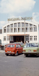 Przed historycznym budynkiem z napisem "Warszawa Główna" zaparkowane są samochody, wśród których dominują pojazdy z lat 70. i 80. XX wieku. Budynek ma symetryczną fasadę z centralnie umieszczonymi drzwiami i oknami po obu stronach. Przestrzeń przed dworcem jest wybrukowana, a na niebie widać lekkie zachmurzenie.