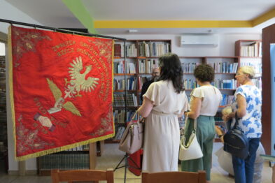 Trzy kobiety oglądają czerwony haftowany baner z białym orłem, który jest zawieszony na stojaku w pomieszczeniu z książkami na półkach. Jedna z kobiet w białej sukience dotyka baneru, jakby badała jego fakturę. Inne osoby stoją wokół i wkładają do torby materiały drukowane.