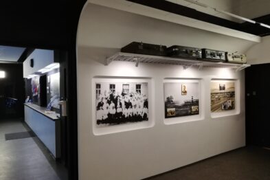 Ściana wystawiennicza z czarno-białymi fotografiami w ramach jest eksponowana w pomieszczeniu o ciemnych ścianach. Na górze ściany ustawiony jest model pociągu na makiecie kolejowej. Widoczne są trzy duże zdjęcia, z których każde przedstawia grupę osób i elementy związane z kolejnictwem.