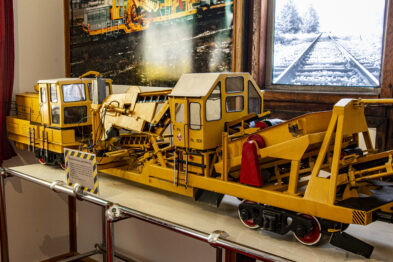 Żółte modele maszyn torowych są wyeksponowane na wystawie; można zauważyć różnorodne pojazdy z wyraźnie widocznymi elementami służącymi do prac na torach. W tle widnieją plakaty ilustrujące tematykę kolejniczą, zwiększając kontekst wystawy dla zwiedzających. Modele są szczegółowe, co pomaga w zrozumieniu ich funkcji w rzeczywistym świecie budowy i konserwacji torów kolejowych.