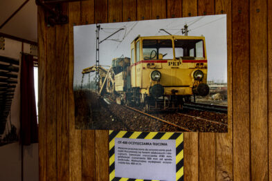 Na zdjęciu widoczny jest plakat przedstawiający żółtą maszynę torową stojącą na torach kolejowych. Maszyna jest wyposażona w koparkę oraz posiada kabiny dla operatorów. Poniżej plakatu umieszczona jest tablica informacyjna z tekstem na żółtym tle.