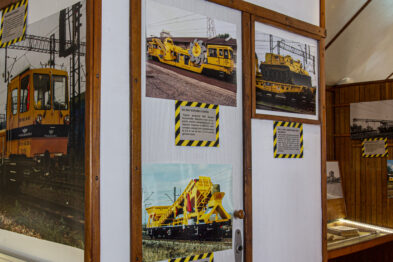 Wewnątrz pomieszczenia wystawienniczego prezentowane są zdjęcia żółtych maszyn torowych na białych ścianach. Fotografie ukazują różne pojazdy używane w pracach kolejowych, takie jak drezyny i podbijarki. Przedstawione maszyny są tłem dla materiałów edukacyjnych, które są wyeksponowane w zasięgu widoku zwiedzających.