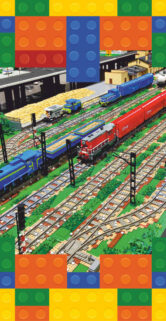 Model kolejowy z klocków Lego ukazuje złożoną scenę z wieloma torami i kolorowymi pociągami. Na jednym z torów widać niebieski lokomotywę z czerwonym wagonem pasażerskim, a obok stoi zielony pociąg towarowy. Otoczenie jest bogate w detale, w tym sygnalizację kolejową, roślinność oraz inne elementy infrastruktury kolejowej.