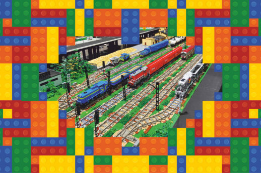Model kolejowy z klocków Lego ukazuje złożoną scenę z wieloma torami i kolorowymi pociągami. Na jednym z torów widać niebieski lokomotywę z czerwonym wagonem pasażerskim, a obok stoi zielony pociąg towarowy. Otoczenie jest bogate w detale, w tym sygnalizację kolejową, roślinność oraz inne elementy infrastruktury kolejowej.
