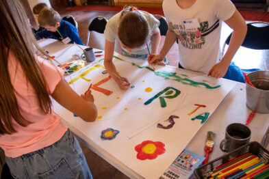 Dzieci używają pędzli i farb do malowania na dużym arkuszu papieru, tworząc kolorowy napis i różnego rodzaju obrazy, takie jak kwiaty. Są one skupione na twórczej pracy, a na stole widać również inne przybory do malowania. Przestrzeń, w której przebywają, jest jasna i przestronna, sprzyjająca kreatywnym działaniom.