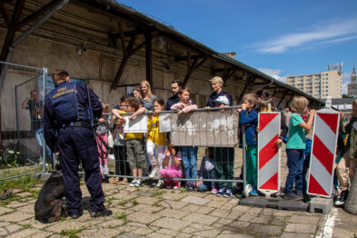 Grupa dzieci stoi na peronie kolejowym obserwując prezentację policjanta w mundurze, który stoi przed nimi z psem. Dzieci są za ogrodzeniem zabezpieczającym, a niektóre z nich unoszą ręce, aby lepiej widzieć. Otoczenie jest nasłonecznione, a w tle widoczne są miejskie budynki.