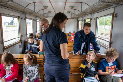 Dzieci siedzą na drewnianych ławkach wewnątrz wagonu kolejowego, skupione na pisaniu i rysowaniu. Dorosła osoba stoi i wskazuje coś dziecku, które jest w stanie z nią współpracować. Wnętrze wagonu jest przestronne, z otwartymi oknami, przez które widać zewnętrzne otoczenie.