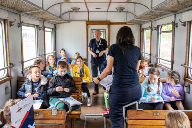 Grupa dzieci siedzi na drewnianych ławkach wewnątrz starego wagonu kolejowego, skupiając uwagę na dorosłej osobie stojącej przed nimi. Osoba ta trzyma otwartą książkę i wydaje się prowadzić zajęcia edukacyjne. W tle widać mężczyznę stojącego i obserwującego interakcję między prowadzącym a dziećmi.