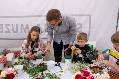 Dzieci w różnym wieku uczestniczą w warsztatach florystycznych pod okiem dorosłego instruktora. Stół, przy którym siedzą, wypełniony jest kolorowymi kwiatami i narzędziami do tworzenia kompozycji kwiatowych. W tle widać baner z napisem 