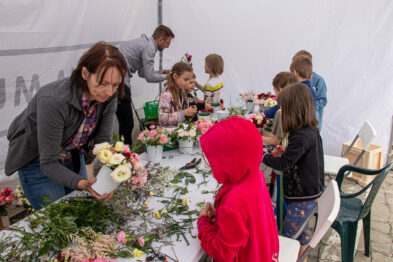 Dzieci w różnym wieku uczestniczą w warsztatach florystycznych, układają bukiety na stołach z kwiatów i różnorodnych roślin. Dorosła osoba pomaga jednemu z dzieci w tworzeniu kompozycji kwiatowej. Na zdjęciu widać także naczynia z kwiatami, zielone gałązki i narzędzia florystyczne rozłożone na stołach.