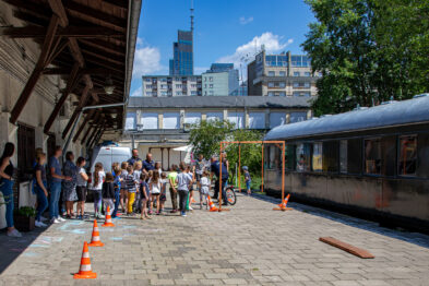 Grupa dzieci uczestniczy w zajęciach na zewnątrz, stojąc obok torów kolejowych i wagonu pociągu. Towarzyszy im kilku dorosłych, a dzieci wydają się słuchać instrukcji. W pobliżu dzieci ustawione są pomarańczowe pachołki tworzące ścieżkę lub tor przeszkód.