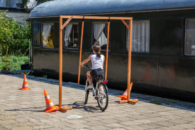 Dziecko na rowerze przemierza tor przeszkód wyznaczony przez pomarańczowe pachołki. Obok toru znajduje się stare, ciemne wagon kolejowy. Słoneczna pogoda i cień wskazują na zewnętrzne, letnie warunki.