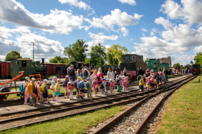 Grupa dzieci kierowana przez dorosłych przemierza teren muzealny wzdłuż torów kolejowych. W tle dostrzegalne są historyczne wagony i lokomotywy. Pogoda sprzyja wydarzeniu, gdyż niebo jest częściowo zachmurzone, a słońce świeci, co widać po cieniach na ziemi.