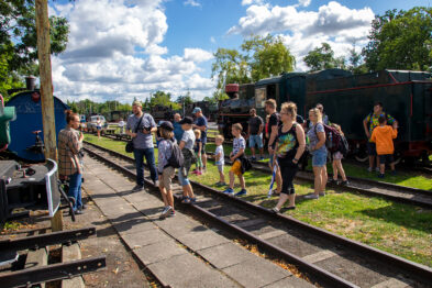 Grupa dzieci pod opieką dorosłych spaceruje po torach kolejowych w muzeum kolejnictwa. Lokomotywy parowe i wagony są zaparkowane po obu stronach torów. Dookoła dominuje zielona trawa i drzewa, a niebo jest częściowo zachmurzone.
