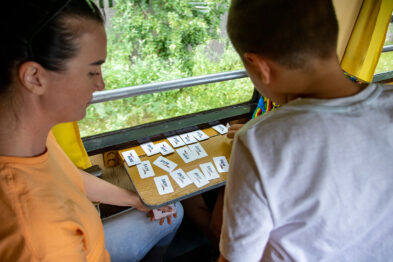 Dwóch uczestników siedzi naprzeciwko siebie przy stoliku wewnątrz wagonu kolejowego, koncentrując się na grze edukacyjnej. Na stole rozłożone są karty, z których dzieci korzystają w trakcie zajęć. Za oknem widoczne są rozmazane zieleń i drzewa, sygnalizujące ruch pociągu.