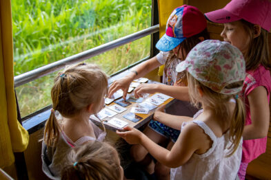 Grupa dzieci siedzi w przedziale kolejowym i skupia się na grze planszowej rozłożonej na białym kartonie. Wszyscy noszą kolorowe letnie kapelusze i wydają się zaabsorbowani działaniem. Poza oknem widać zieloną roślinność, co sugeruje, że pociąg znajduje się w ruchu lub na stacji otoczonej zielenią.