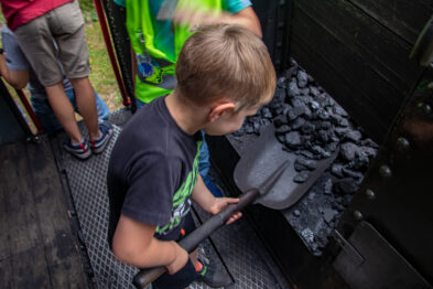Dziecko trzyma dużą, metalową łopatę i przekłada nią czarne kawałki węgla w przestrzeni, która przypomina tendrzak parowozu. Jego skupienie sugeruje zaangażowanie w działanie, które wykonuje. W tle widoczni są inni uczestnicy zajęć, rozmyci i częściowo widoczni zza parapetu.
