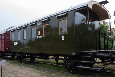 Odstawiony zielony wagon kolejowy stoi na torach; widoczne są jego drewniane elementy i drzwi z oknami. Kilka informacyjnych plakatów jest wywieszonych na zewnątrz wagonu, na którym czytelne są litery 