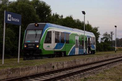 Nowoczesny pociąg w kolorach zielono-niebieskich stoi na stacji kolejowej Basznia obok której wisi tabliczka z nazwą stacji. Za pojazdem znajdują się częściowo zarośnięte trawniki i kilka drzew, a w tle widać niebieskie niebo z chmurami w blasku zachodzącego słońca. Tor kolejowy rozciąga się równolegle do pociągu, biegnąc w kierunku horyzontu.
