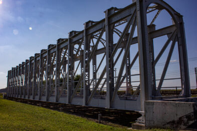 Metalowy most kolejowy o konstrukcji przęsłowej znajduje się na otwartym terenie pod błękitnym niebem. Na pierwszym planie widać solidne betonowe podpory, na których opiera się stalowa konstrukcja mostu złożona z wielu równoległych łuków. Tor kolejowy przechodzi przez most, a jego bieg wiedzie w kierunku niezamieszkałych obszarów.