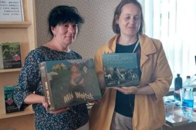 Dwie kobiety stoją obok siebie w pomieszczeniu z książkami i trzymają egzemplarze albumu 